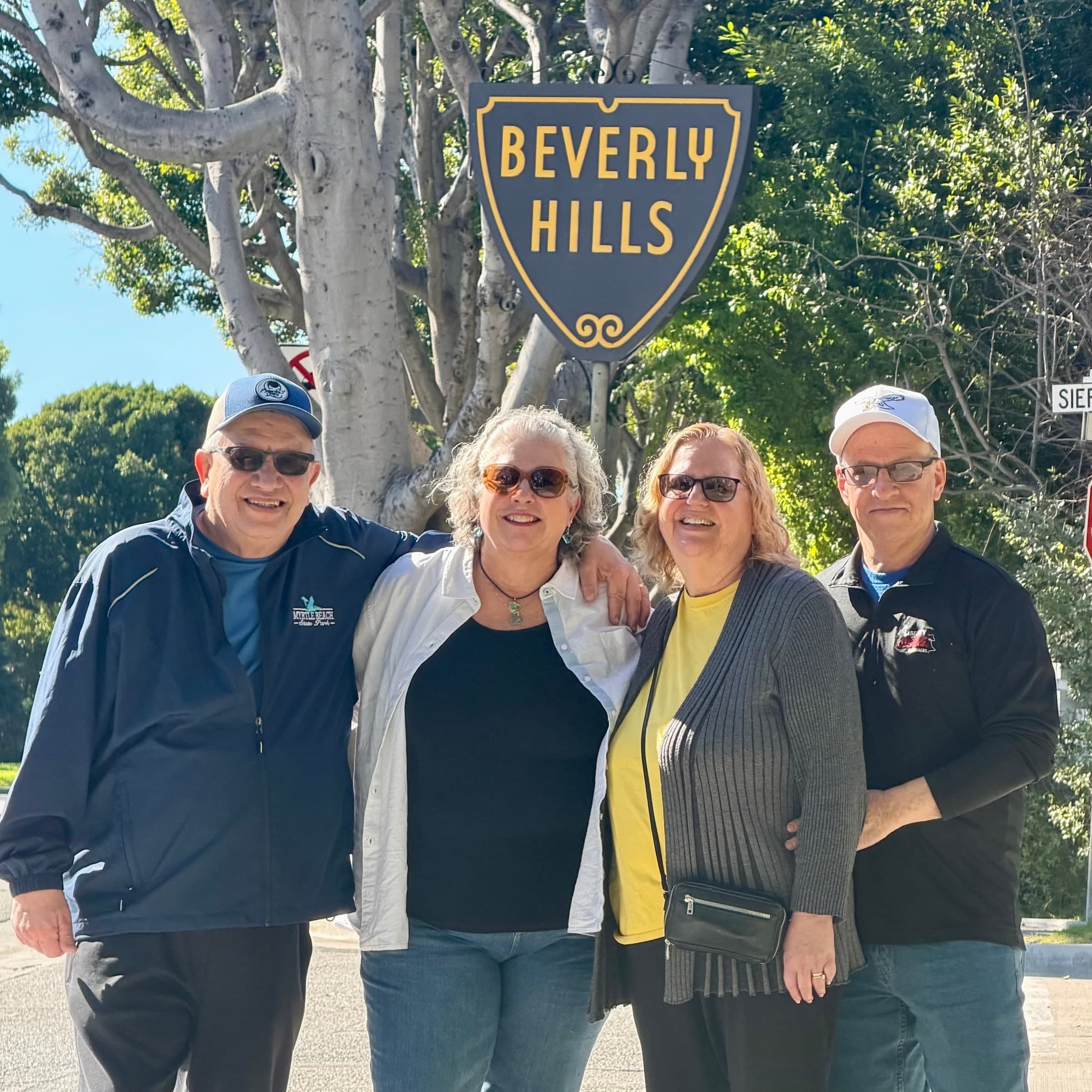 Four guests smiling beside the Beverly Hills shield sign under a tree