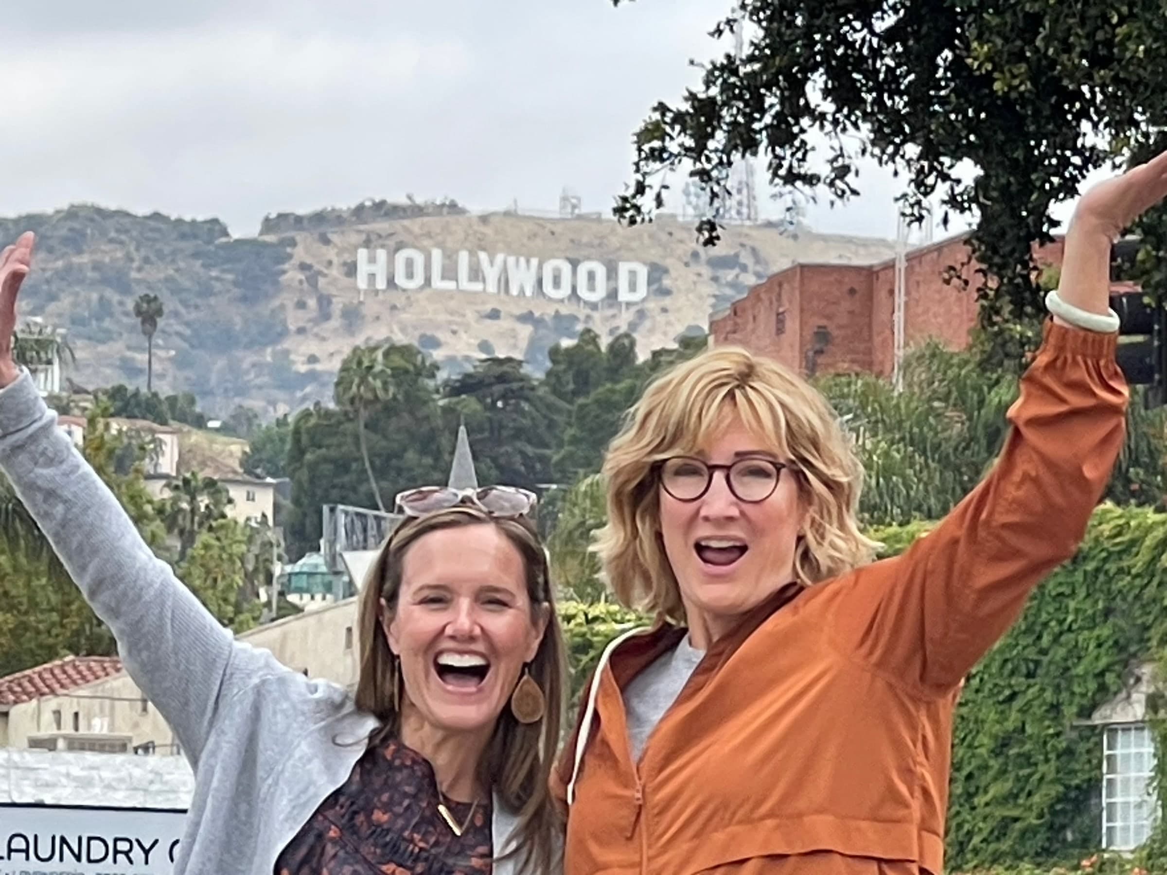 Two women cheering with their arms raised, the Hollywood Sign on the hills behind them