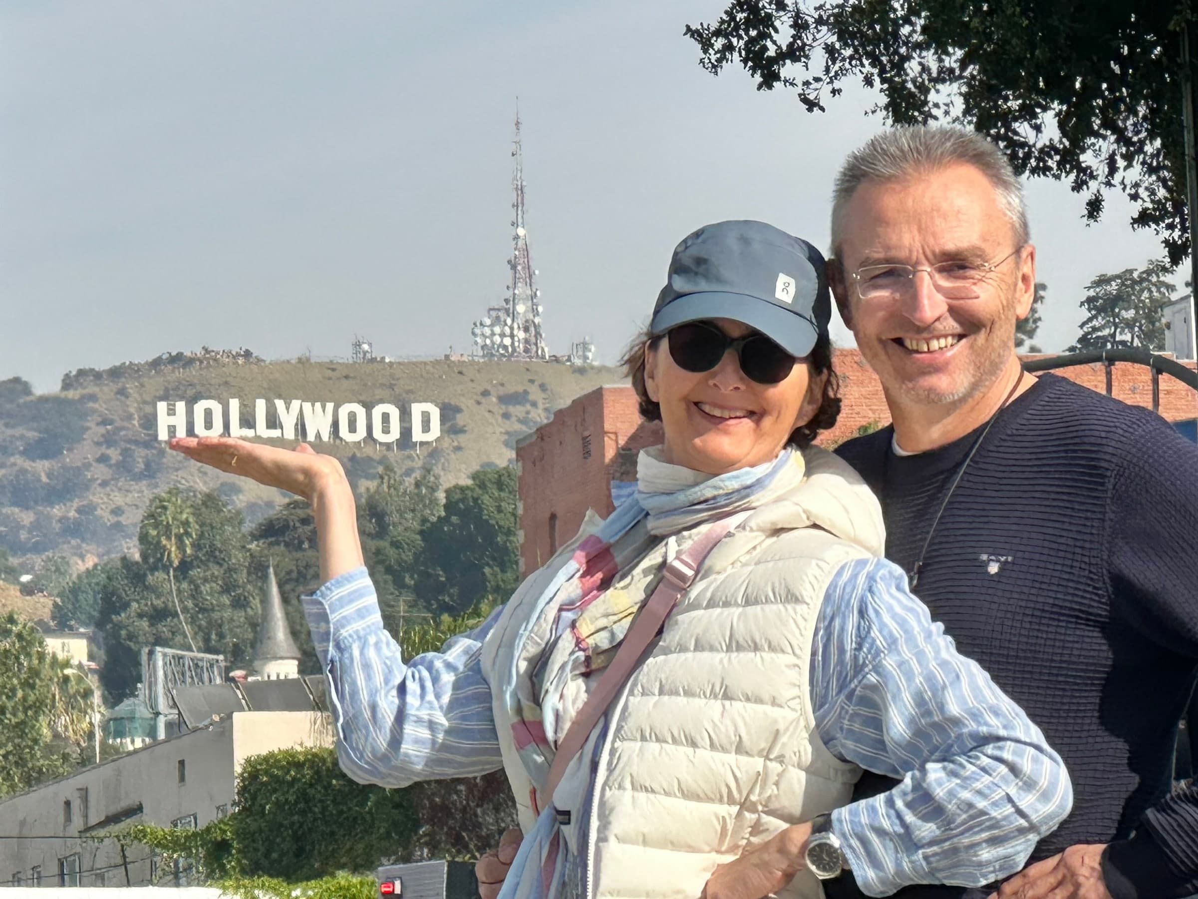 A couple smiling, she holds out her hand toward the Hollywood Sign