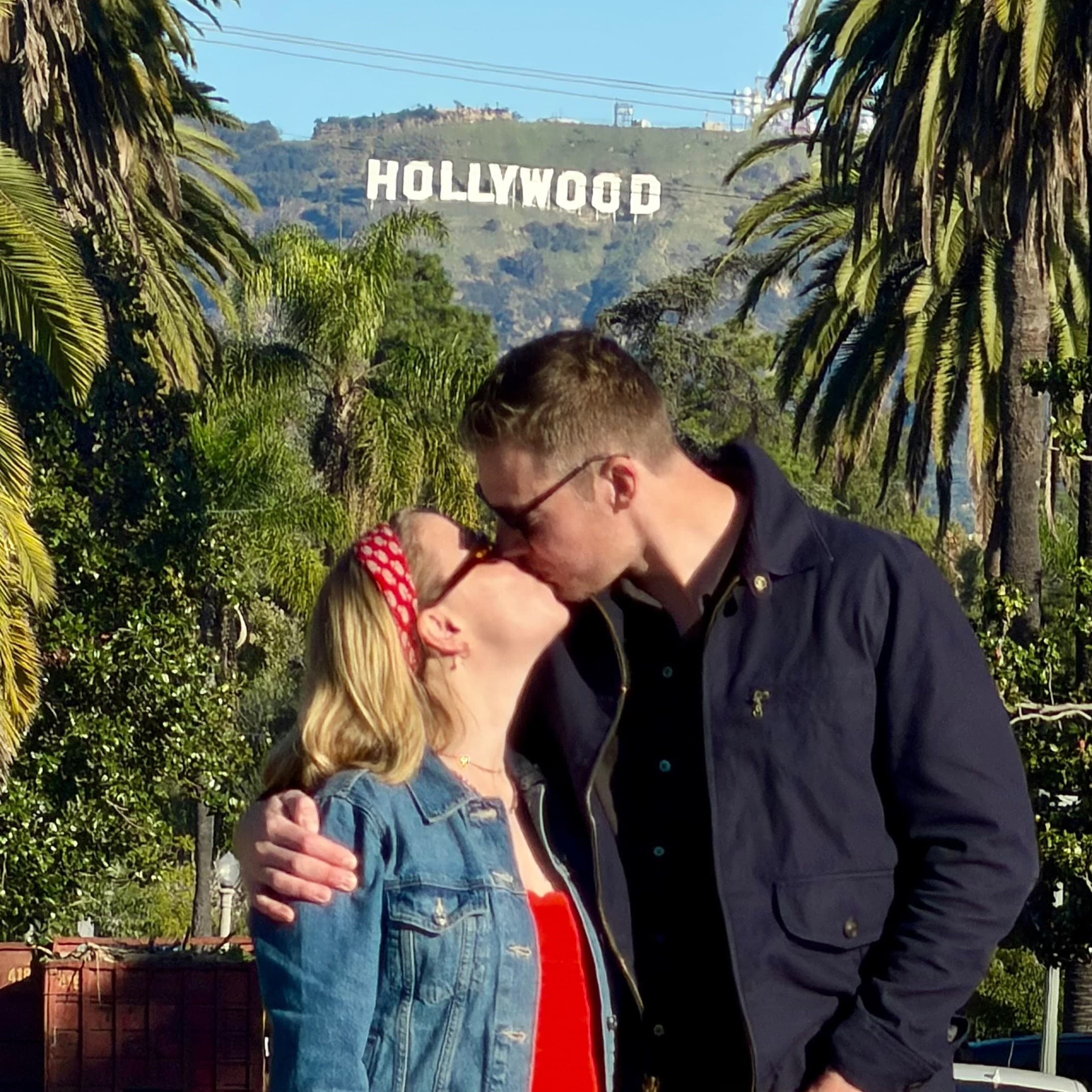 A couple kissing among palm trees with the Hollywood Sign behind them