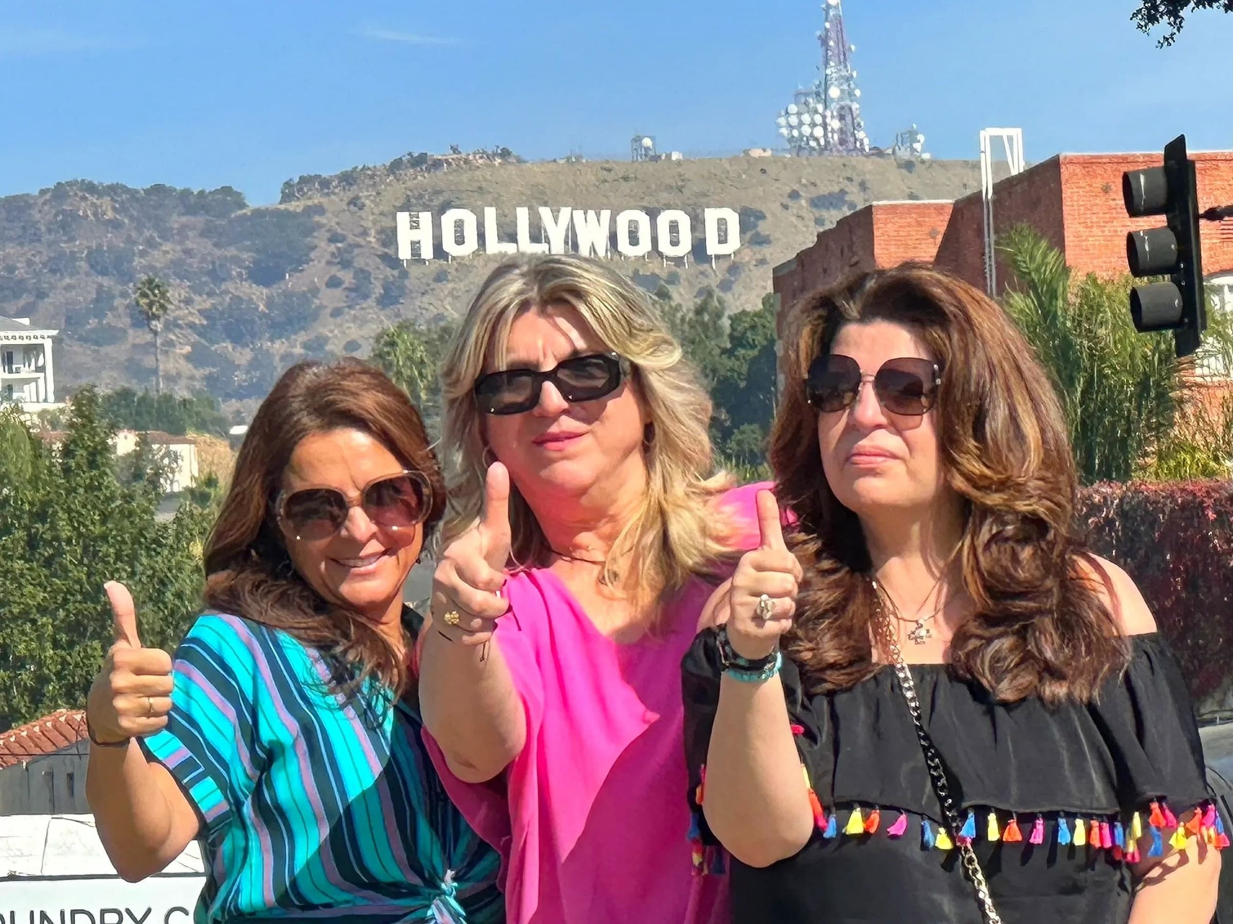 Three women in sunglasses giving a thumbs up below the Hollywood Sign