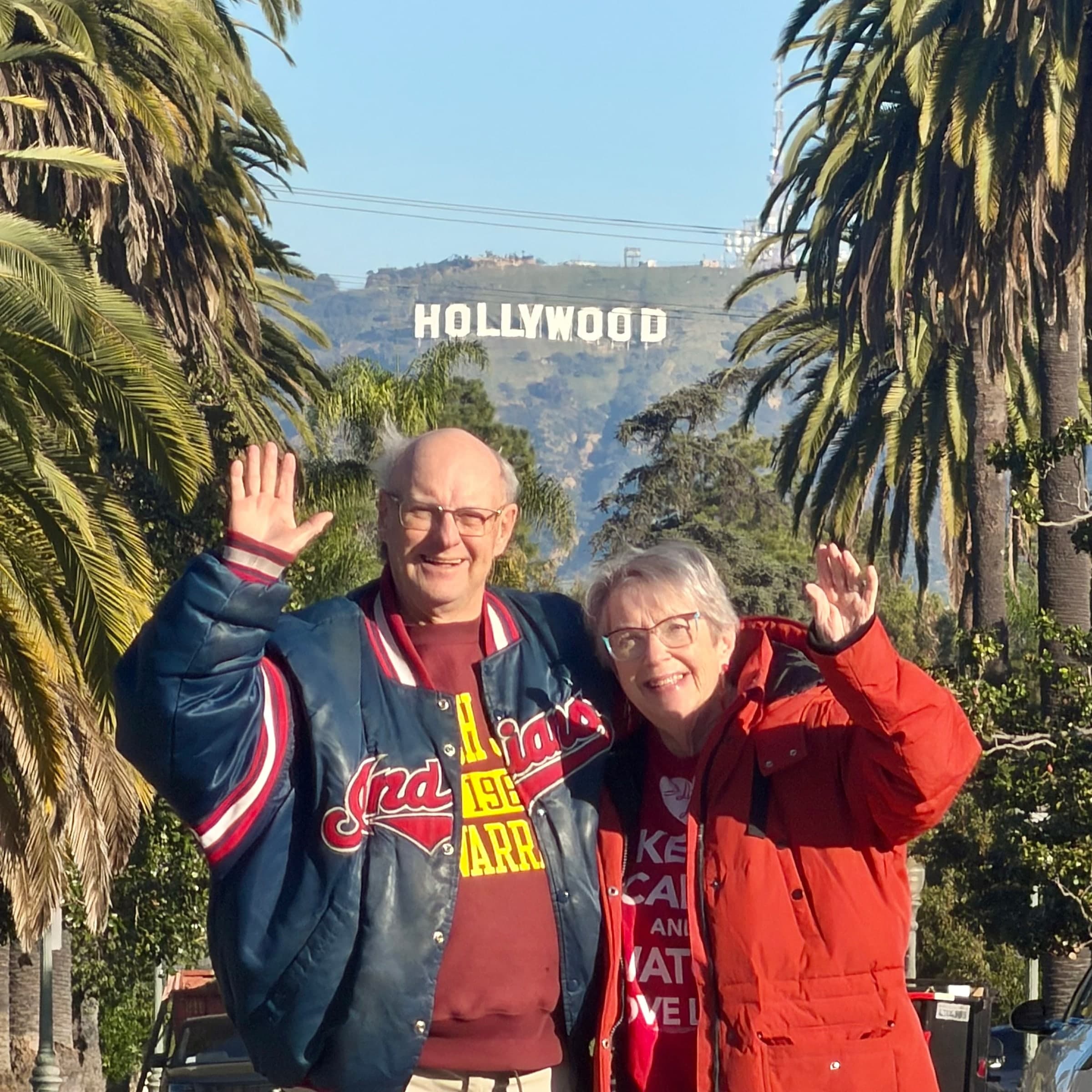 An older couple waving in front of palm trees, the Hollywood Sign on the hill
